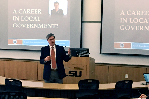 Man speaks in front of classroom with podium and screens in background. 